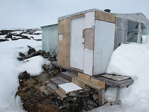 The entrance to a cabin where the snow seems to have melted; it’s thick snow all around. A staircase of boards is placed on packed stones. The cabin is an assembly of pieces of wood and sheet metal; some are covered with faded pale paint.