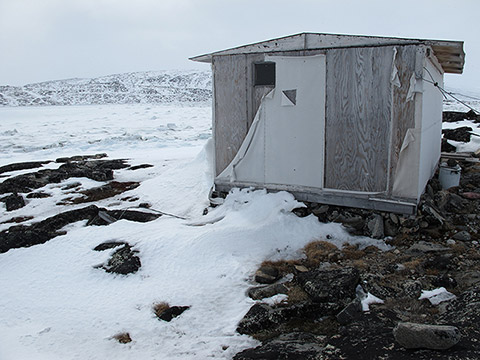 On the rocky and partially snow-covered shores of a frozen creek, a cabin rests on a pile of stones. It’s made of used wooden panels. Certain pieces, torn and hanging, come from an old canvas covering.